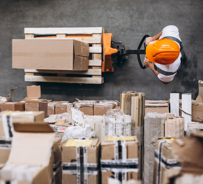 Worker in orange safety helmet operating a manual pallet jack to move a pallet of cardboard boxes in a warehouse, surrounded by organized stacks of packages and materials.